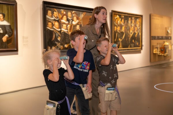 drie kinderen staan naar kunst te kijken met een verrekijker. hun moeder staat achter hen en kijkt ook.